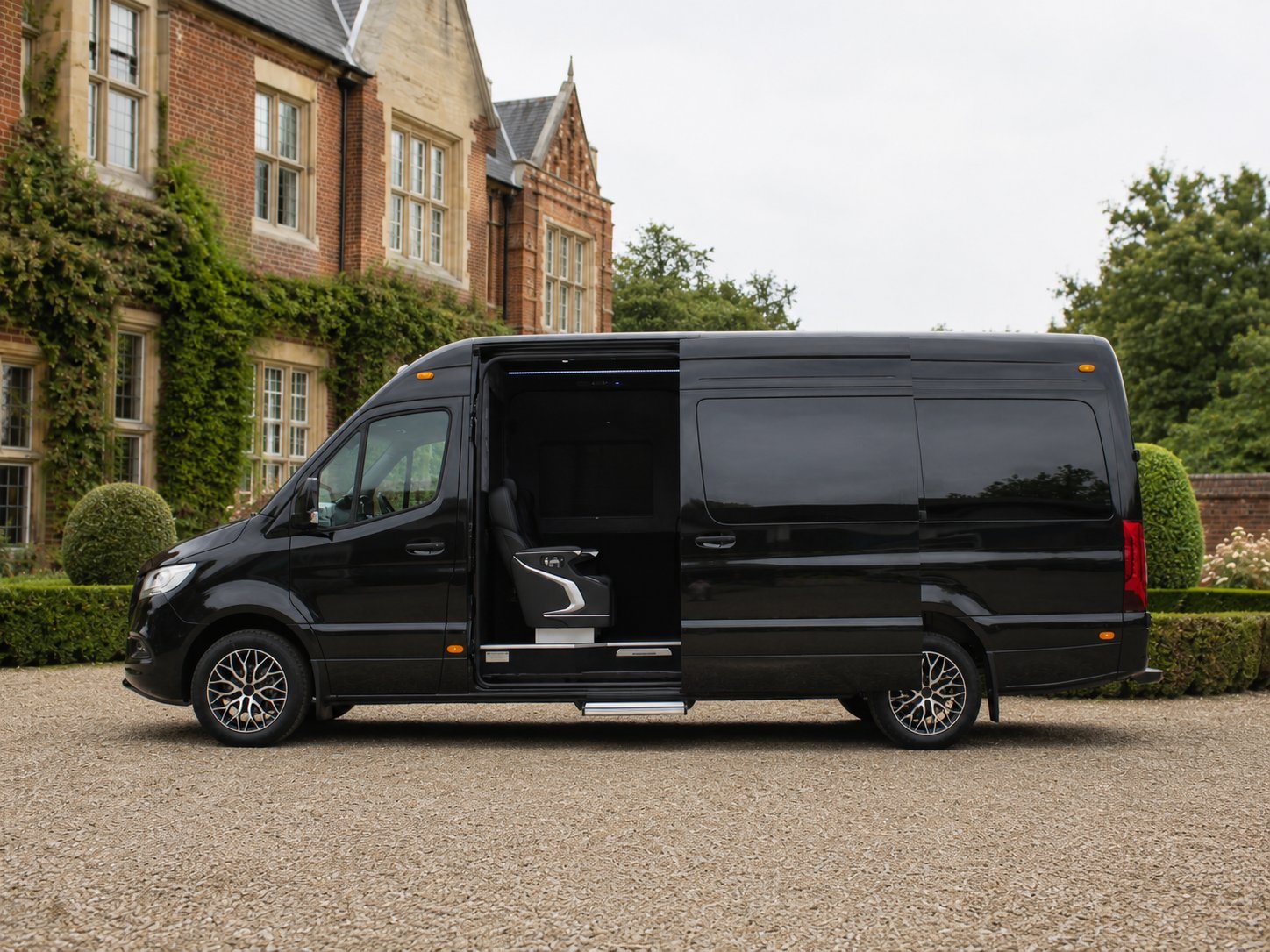 Black luxury shuttle van with open sliding door parked in front of brick mansion with ivy-covered walls