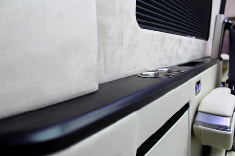 Interior of an RV showing a bed with white bedding and dark trim along the window area