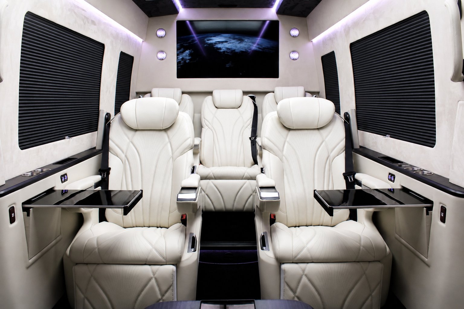 Luxury van interior with white leather captain seats, mounted displays, dark window shades, and ambient lighting