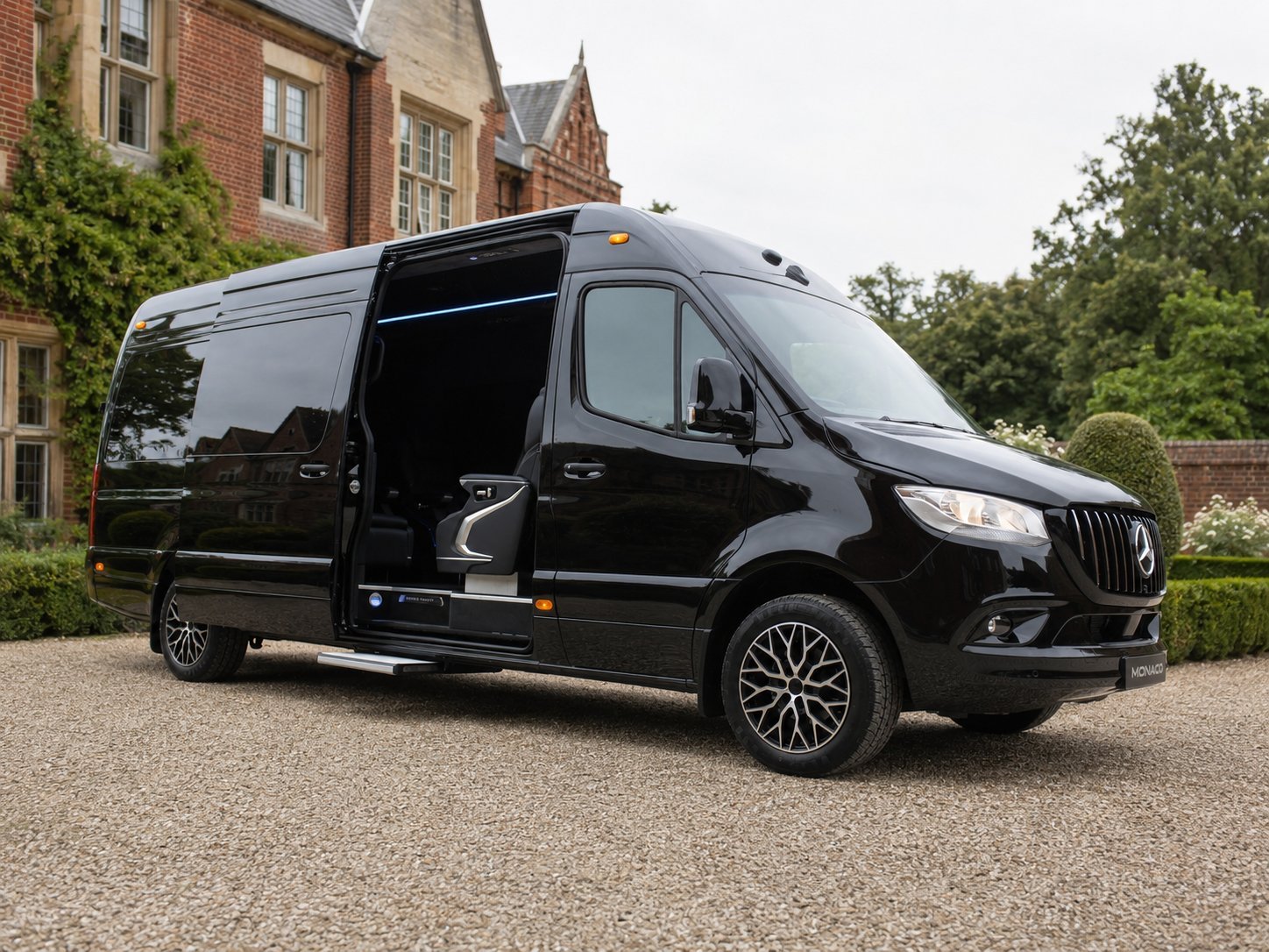 Black luxury minivan parked on gravel driveway in front of brick mansion with ivy and trees