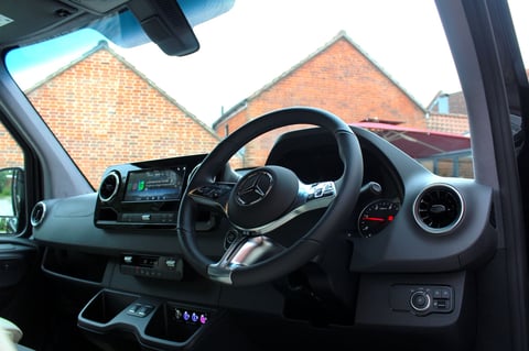 Interior view of Mercedes vehicle dashboard showing steering wheel, digital display, and brick buildings visible through windshield