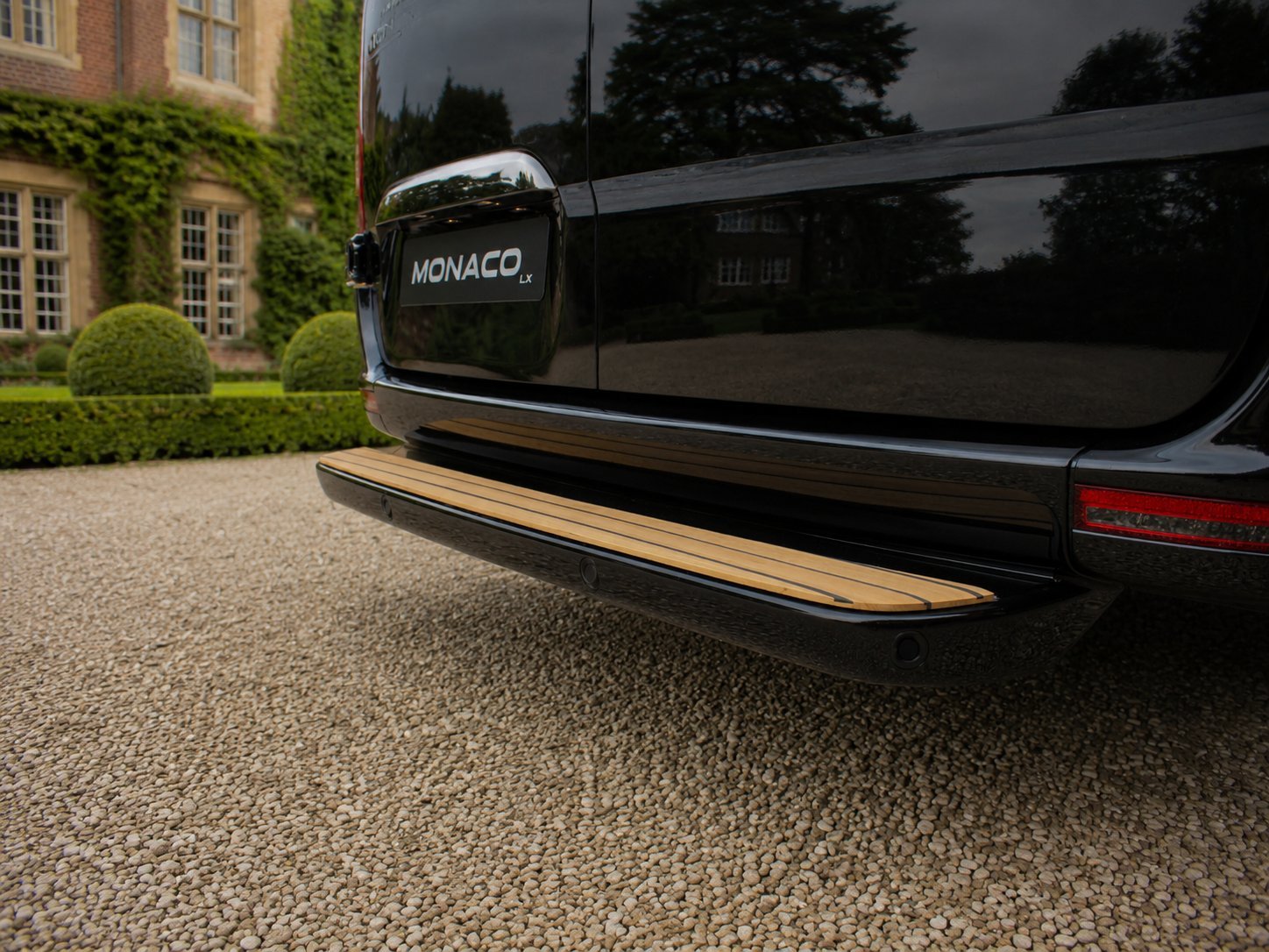 Rear view of black Monaco luxury vehicle parked on gravel driveway with ivy-covered building and manicured garden in background