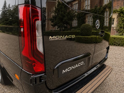 Rear view of a black Mercedes-Benz Monaco van showing red taillights and badging on a gravel driveway
