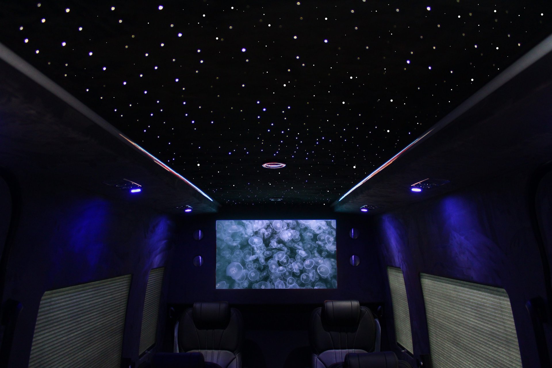 Modern home theater with starry ceiling, blue ambient lighting, wall speakers, and large center screen displaying celestial imagery