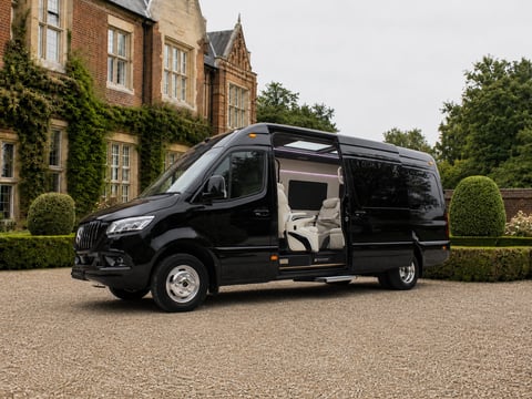 Black luxury van parked in front of a brick mansion with open side door, surrounded by manicured hedges