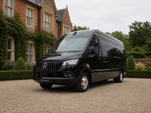 Black luxury minibus parked on gravel driveway in front of brick mansion with ivy-covered walls
