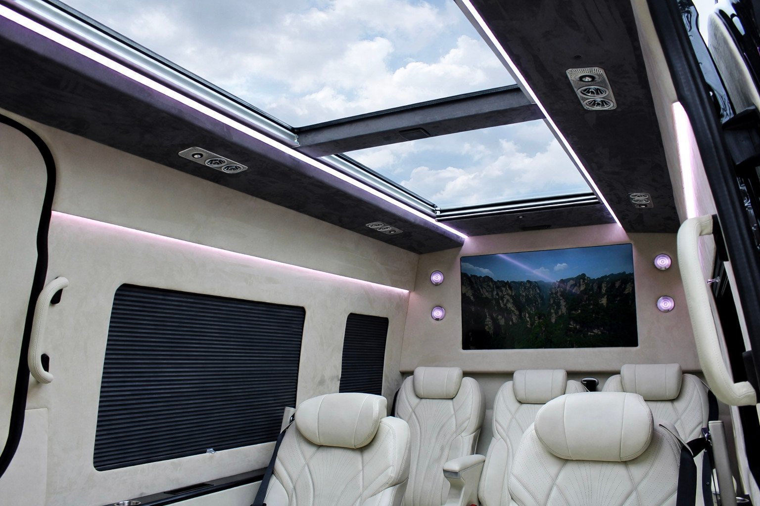 Luxury RV interior with panoramic sunroof, cream-colored seating, mounted TV screen, and forest mountain view displayed on screen