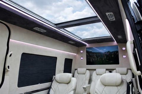 Luxury RV interior with panoramic sunroof, cream-colored seating, mounted TV screen, and forest mountain view displayed on screen